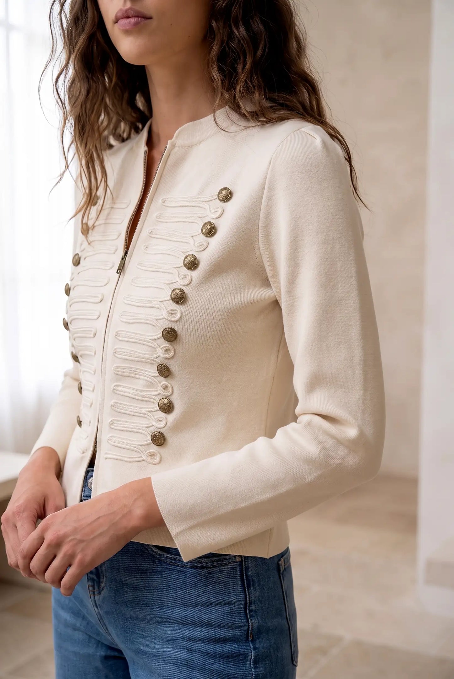 Chaqueta corta color crema con botones dorados y detalles de cordones frontales.