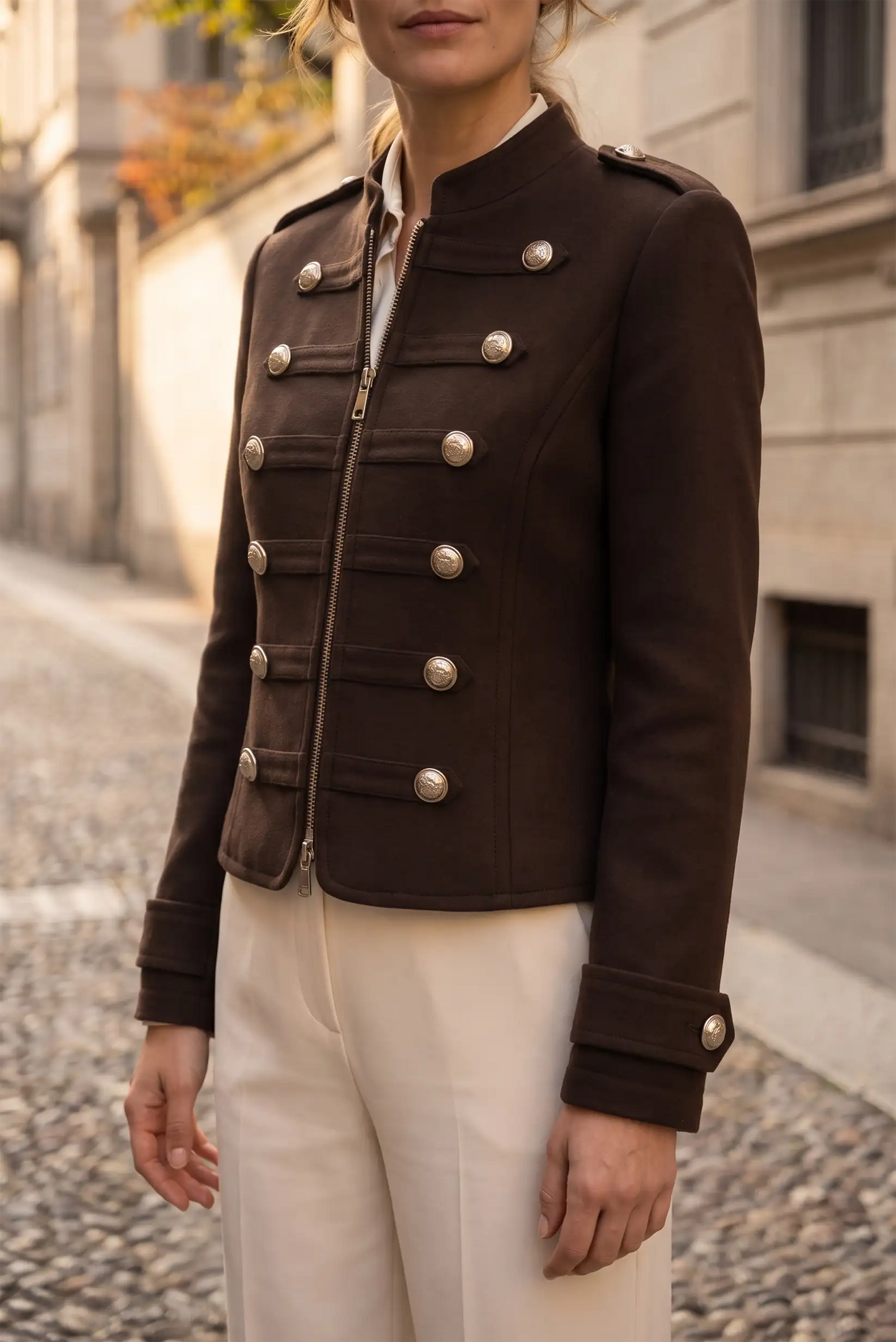 Mujer vistiendo chaqueta marrón estilo militar con botones plateados y pantalones blancos