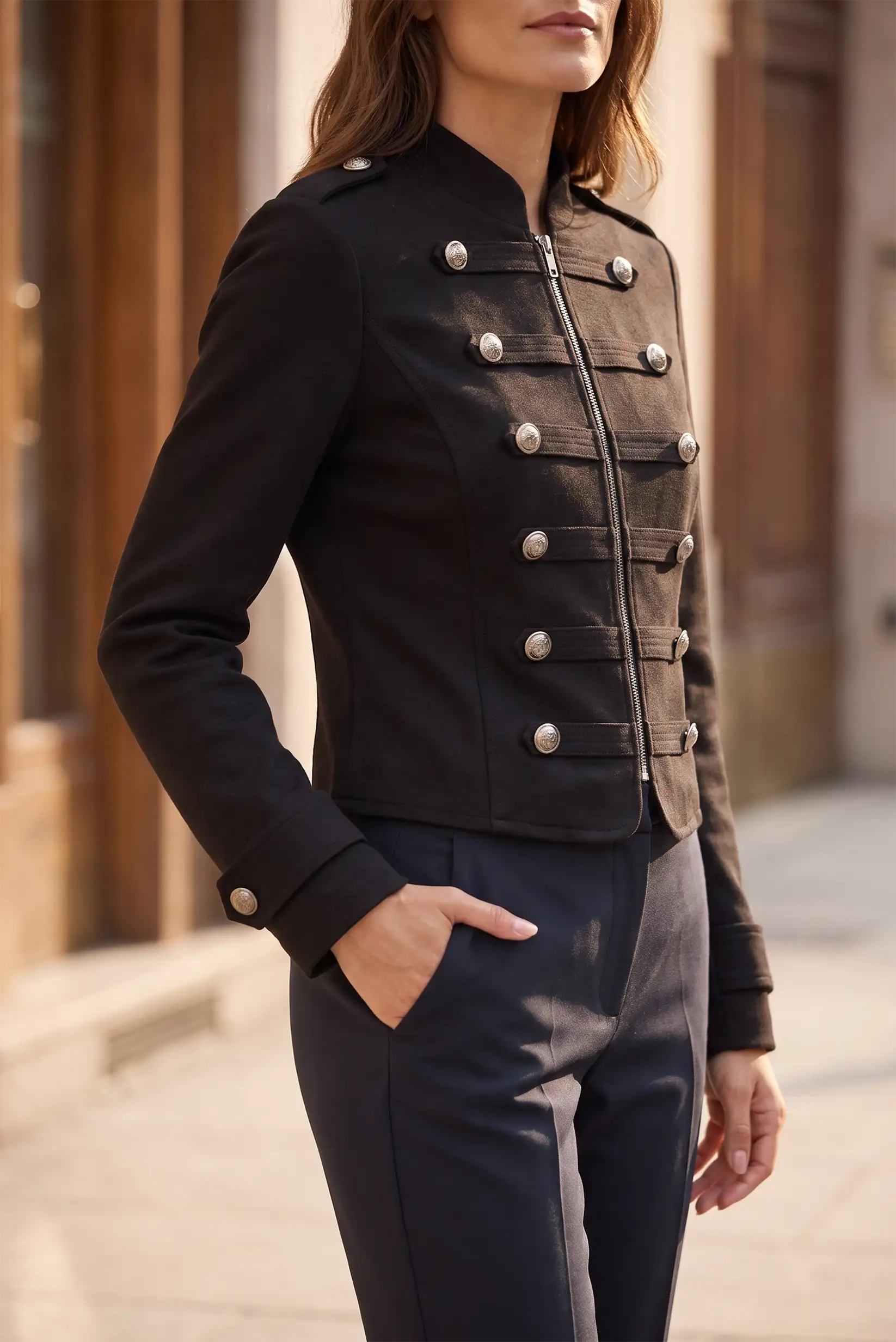 Mujer vistiendo chaqueta militar negra corta con botones plateados y pantalón oscuro.