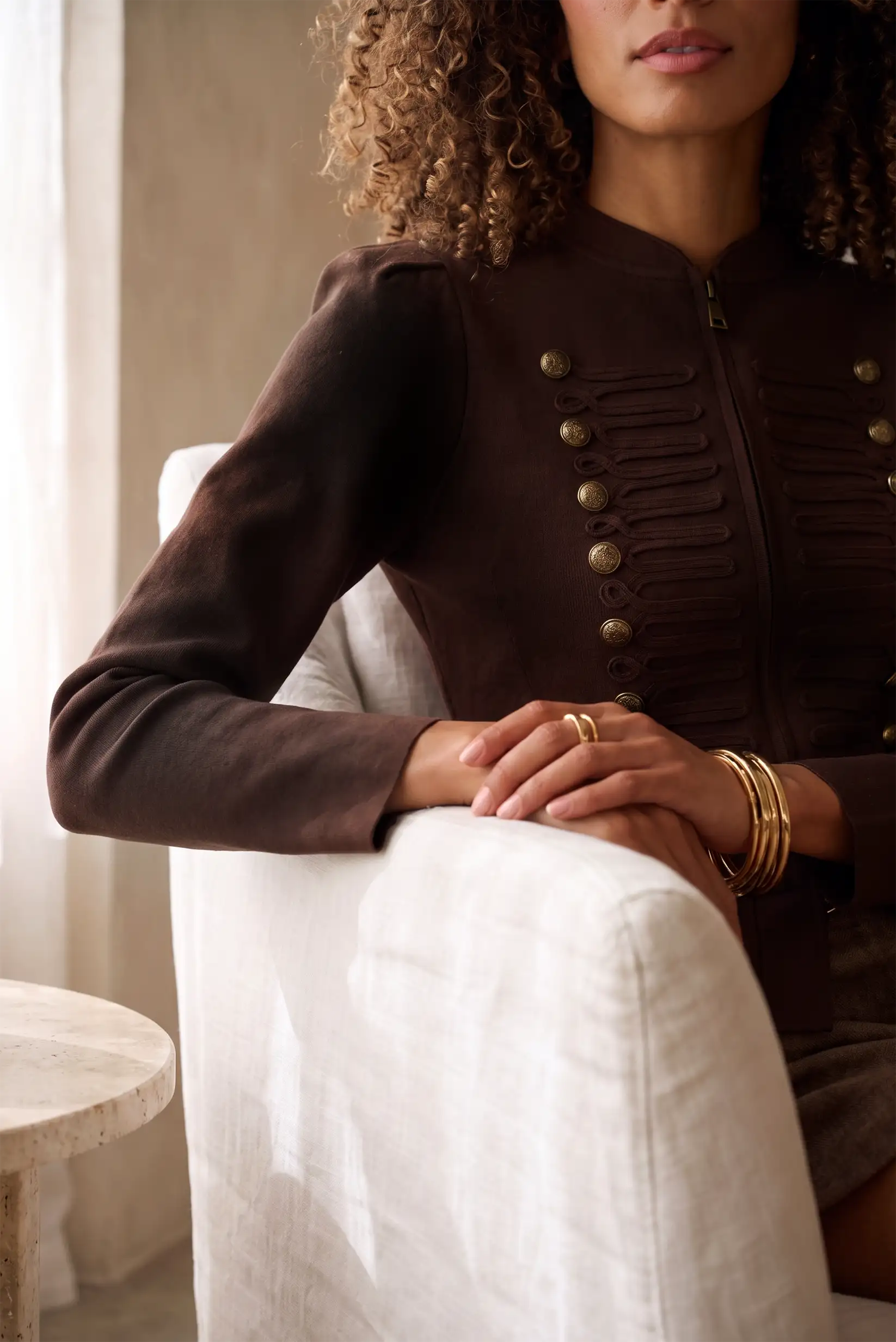 Mujer vistiendo chaqueta marrón estilo militar con botones dorados y detalles plisados.