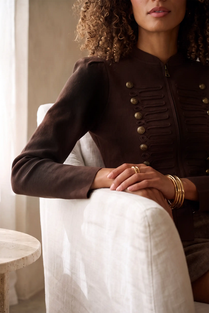 Mujer vistiendo chaqueta marrón estilo militar con botones dorados y detalles plisados.