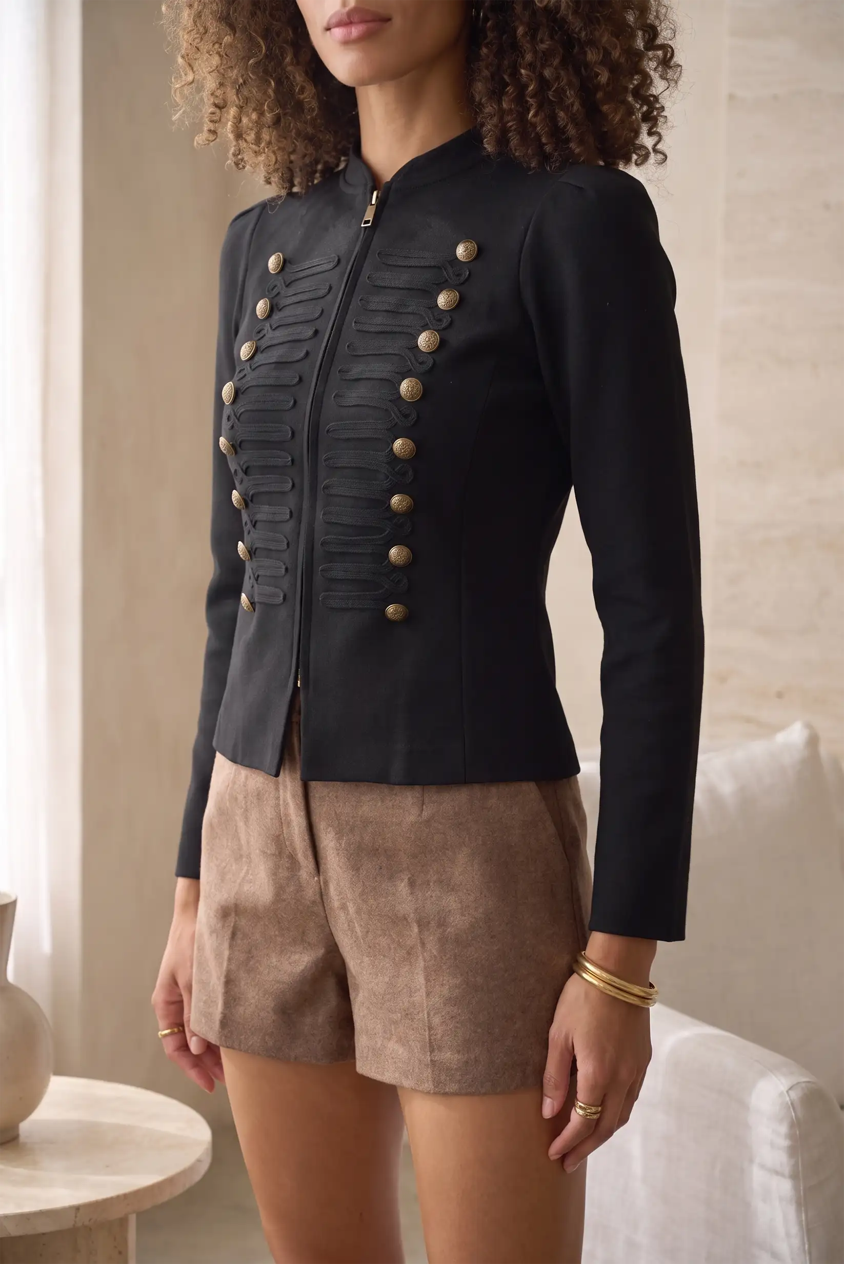 Mujer vistiendo chaqueta negra estilo militar con botones dorados y shorts beige.