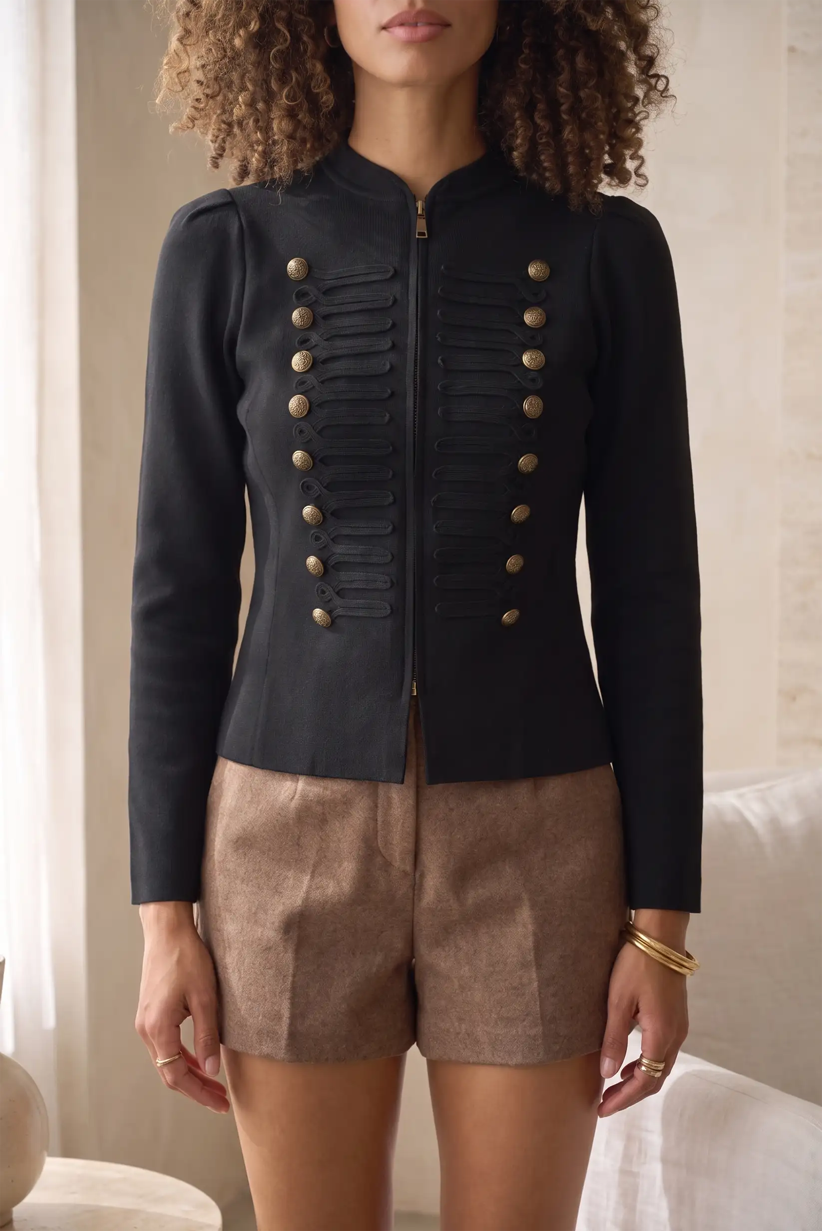 Mujer vistiendo chaqueta negra estilo militar con botones dorados y shorts marrones.