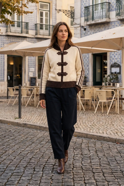 Mujer con chaqueta beige y marrón con detalles bordados y pantalón negro en calle empedrada.