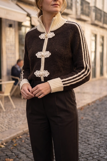 Mujer con chaqueta de punto marrón con detalles blancos y pantalones oscuros en calle empedrada.