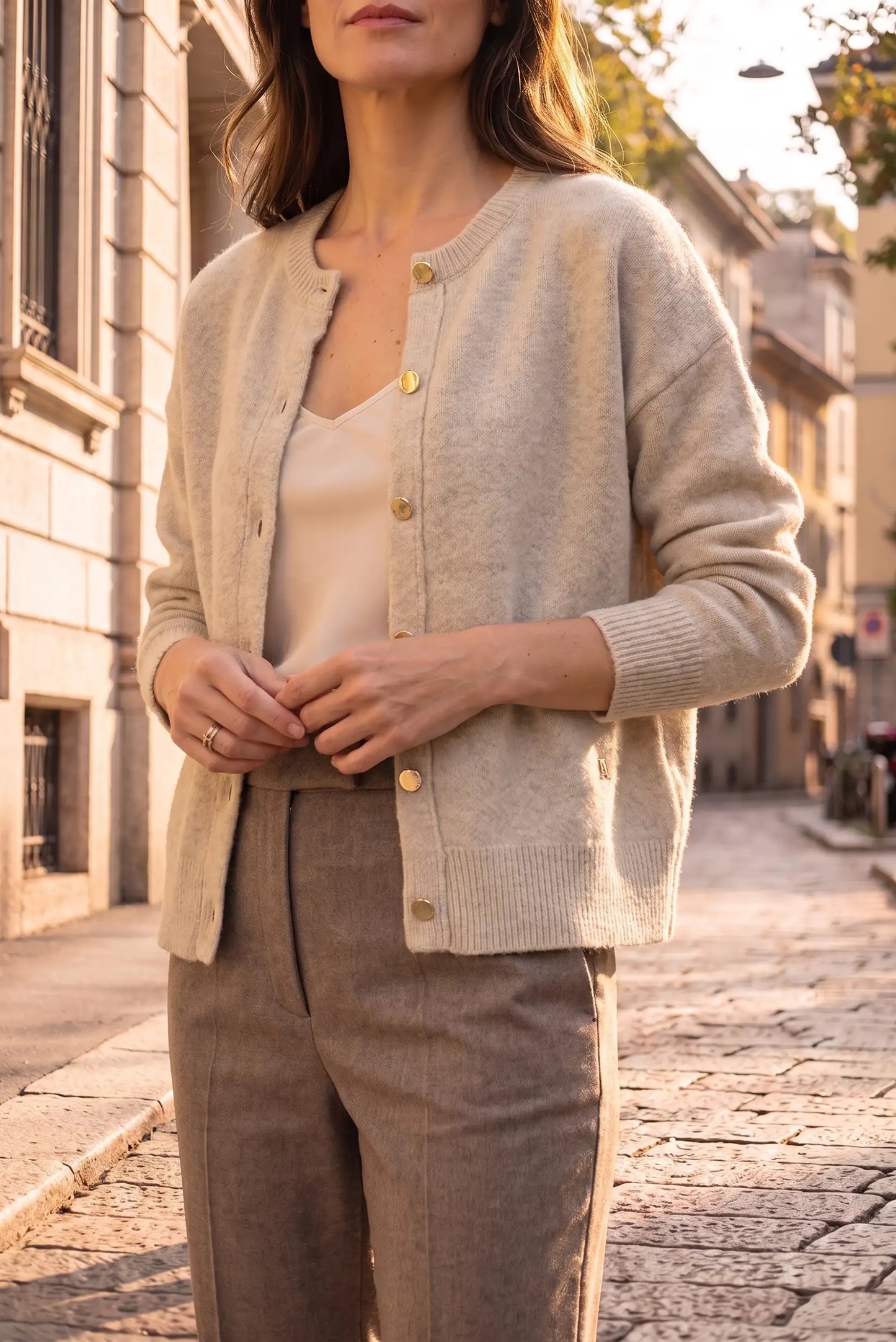 Mujer vistiendo rebeca de punto beige con botones dorados y pantalones marrones en calle urbana.
