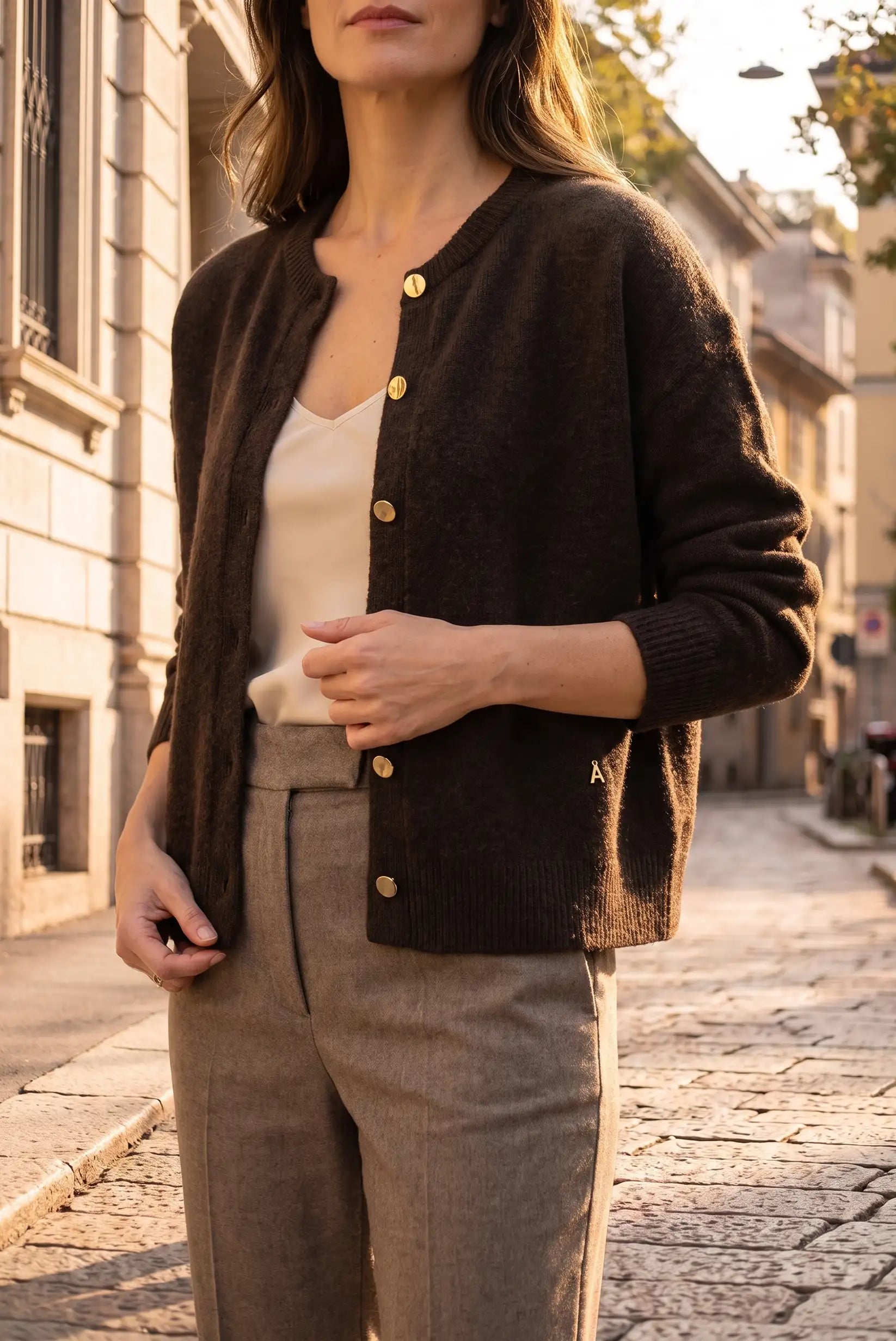 Mujer vistiendo cárdigan marrón oscuro con botones dorados y pantalones beige en calle urbana.