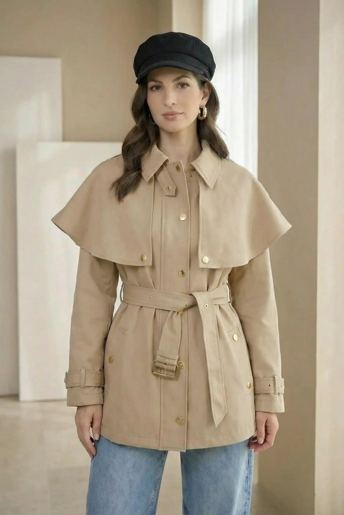 Mujer con chaqueta trench beige, cinturón y botones dorados, jeans y gorra negra.
