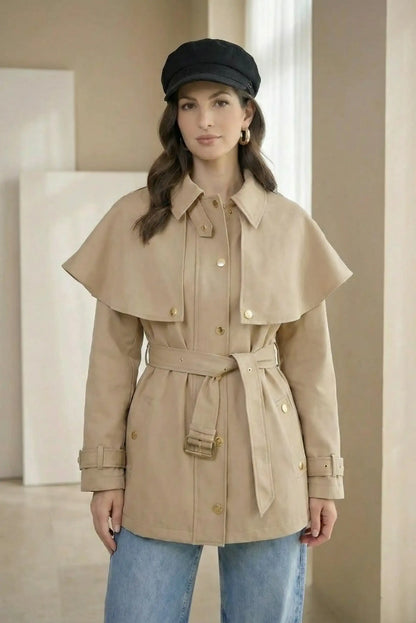 Mujer con chaqueta trench beige, cinturón y botones dorados, jeans y gorra negra.