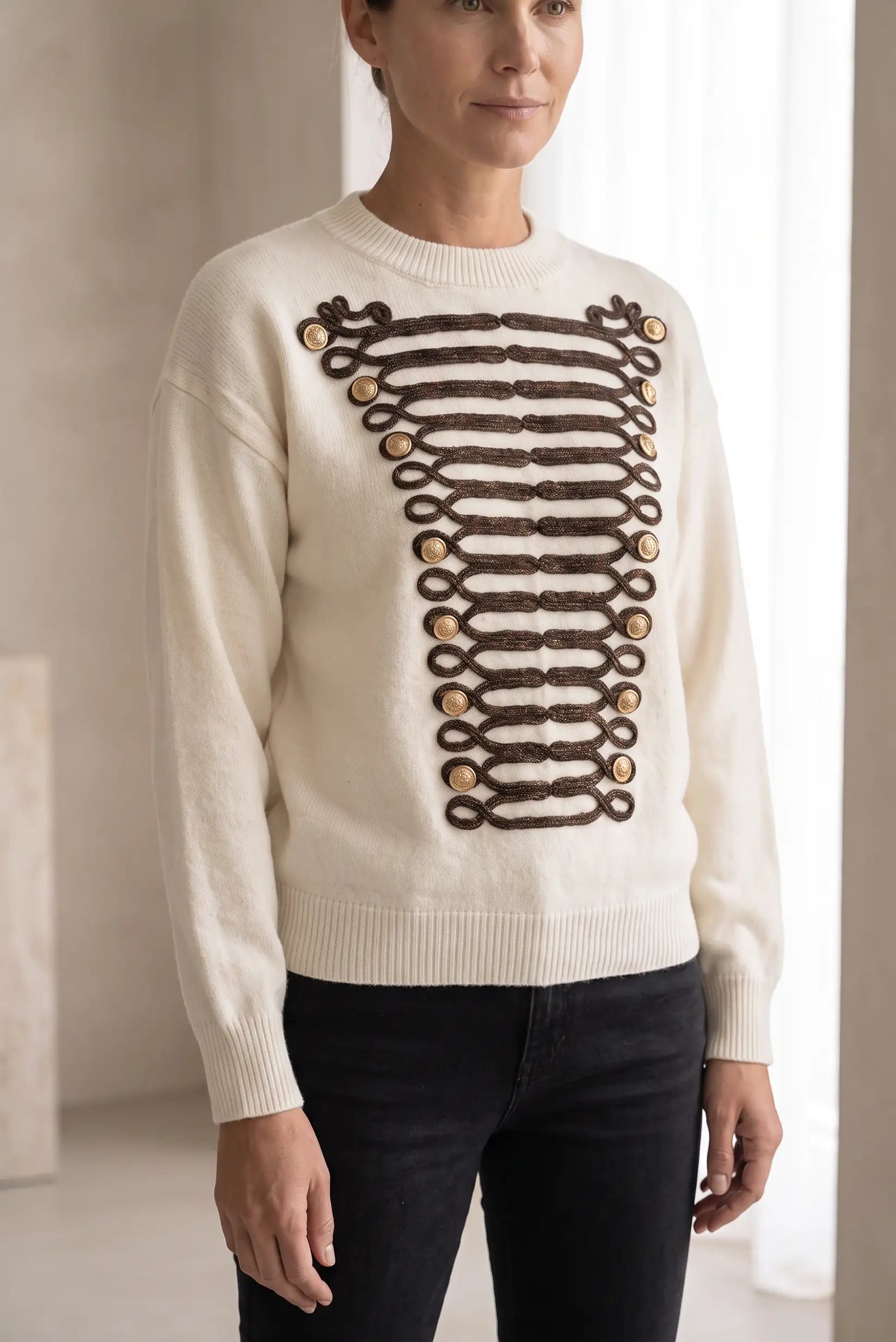 Mujer vistiendo jersey blanco con detalles decorativos de botones dorados y cordones marrones.
