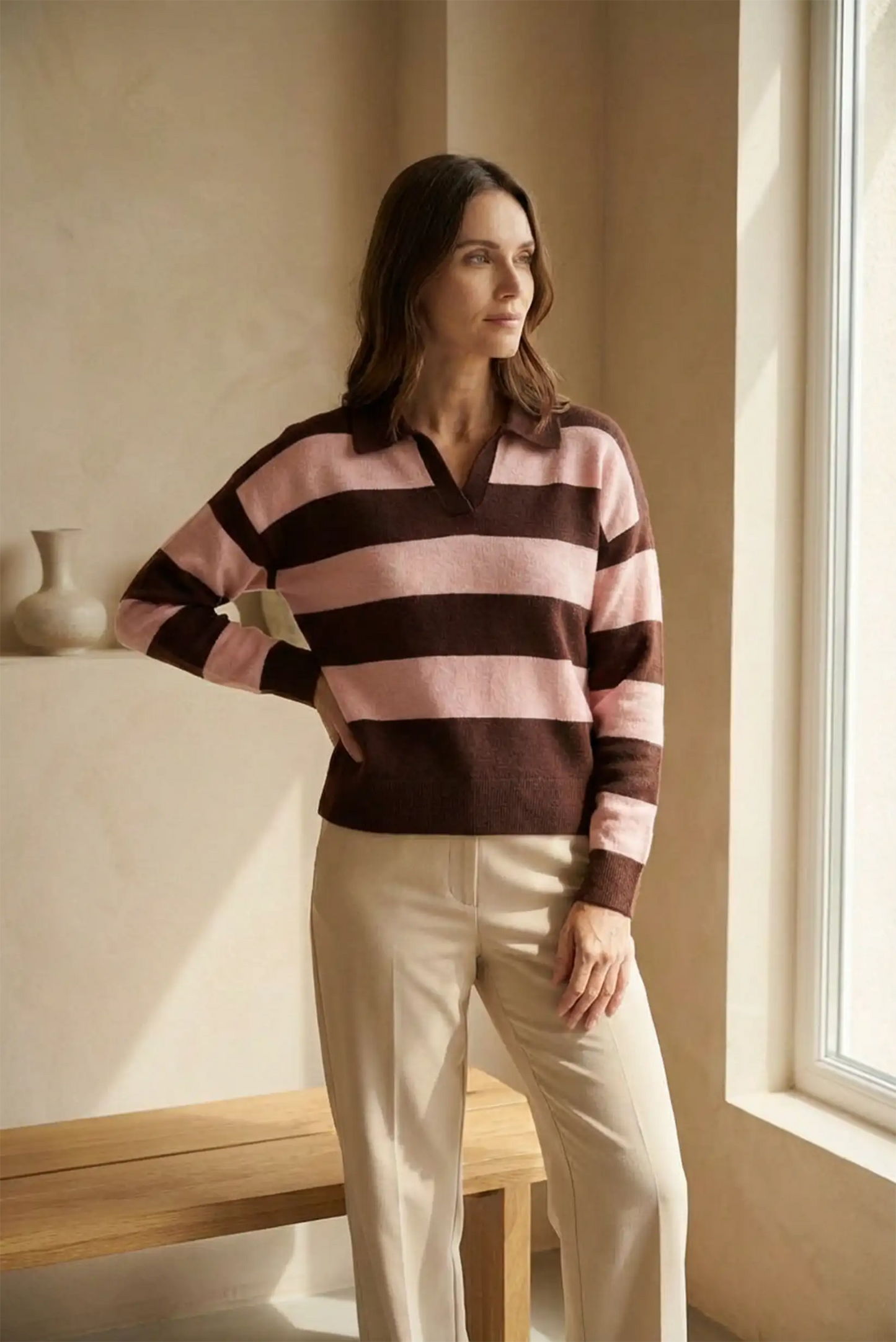 Mujer con jersey de manga larga rayas rosa y marrón, pantalones beige, junto a ventana y banco de madera