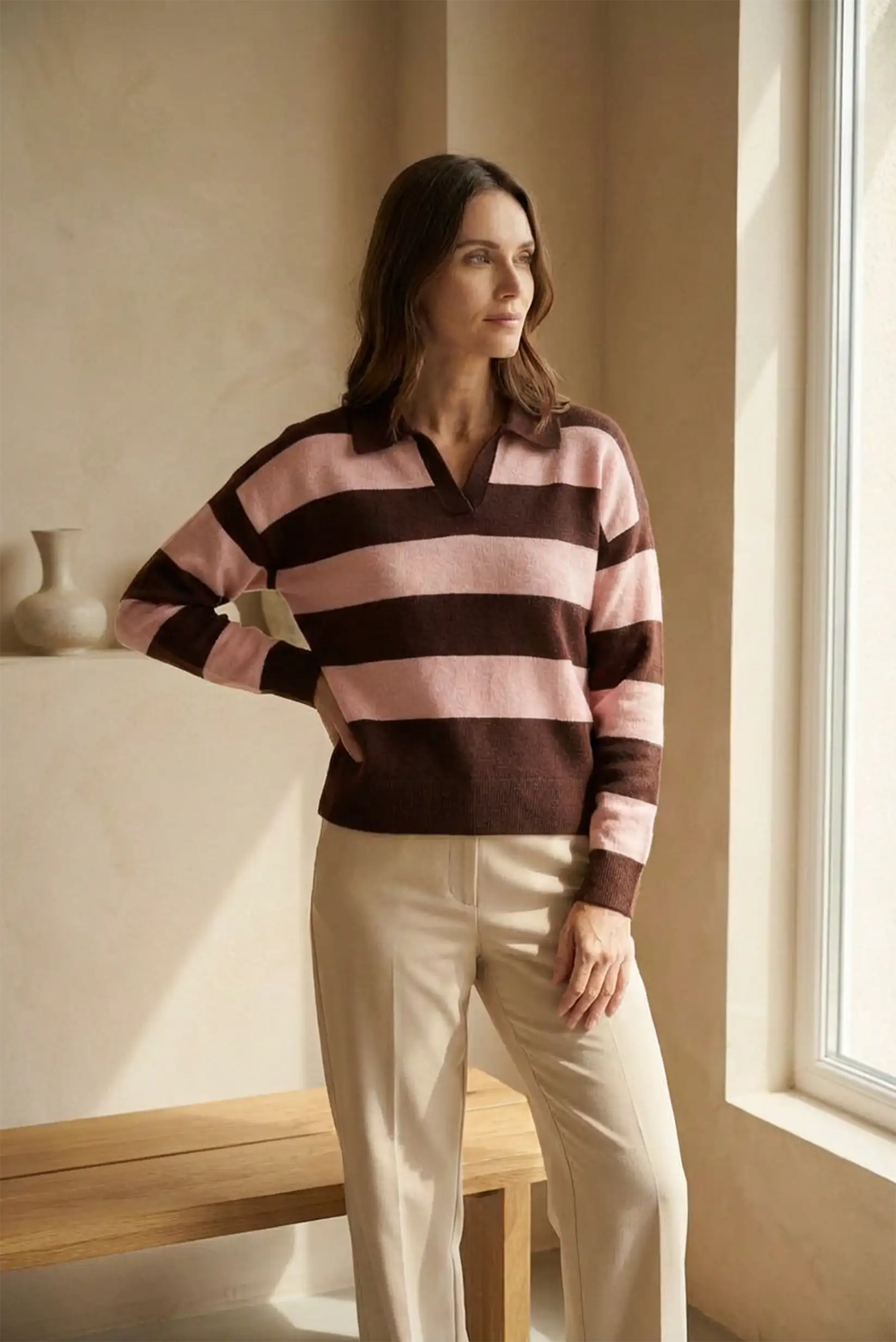 Mujer con jersey de manga larga rayas rosa y marrón, pantalones beige, junto a ventana y banco de madera