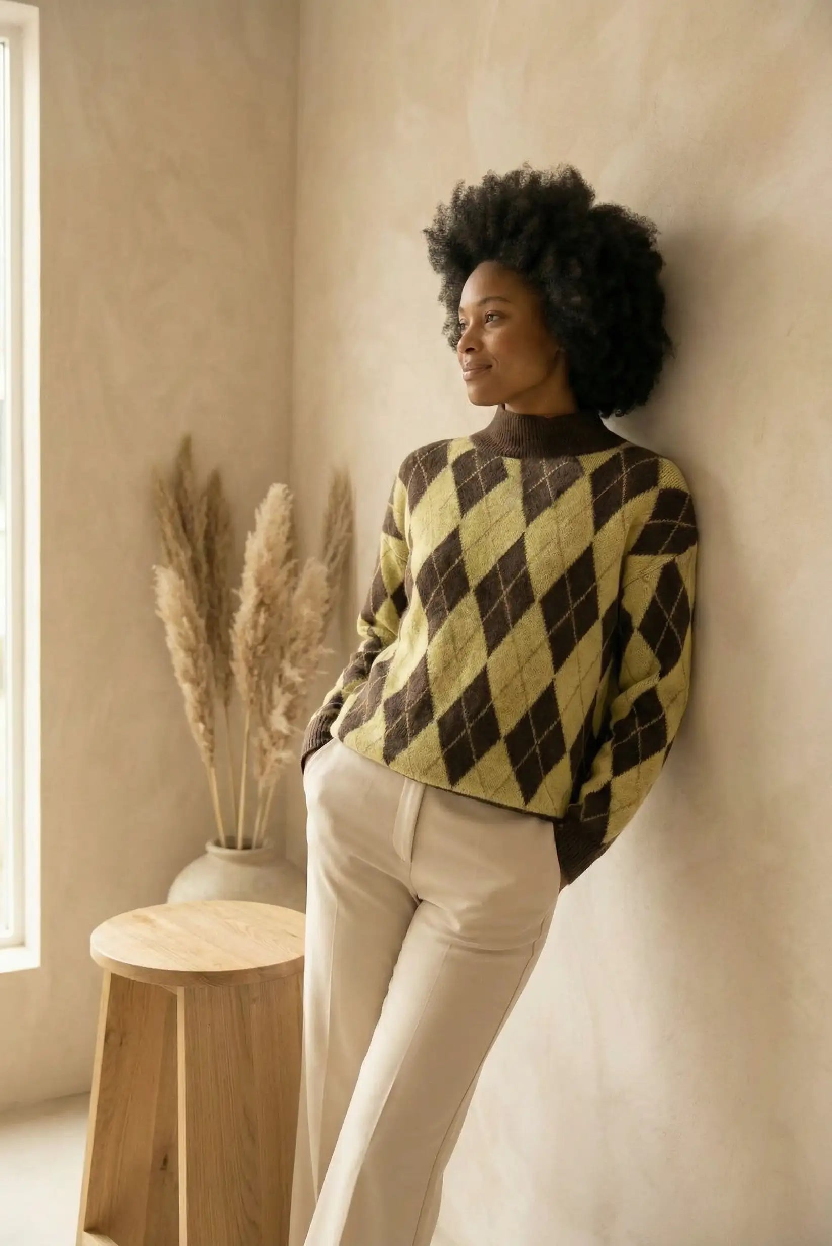 Mujer con jersey de rombos amarillo y marrón, pantalones beige, en interior minimalista con jarrón y taburete de madera