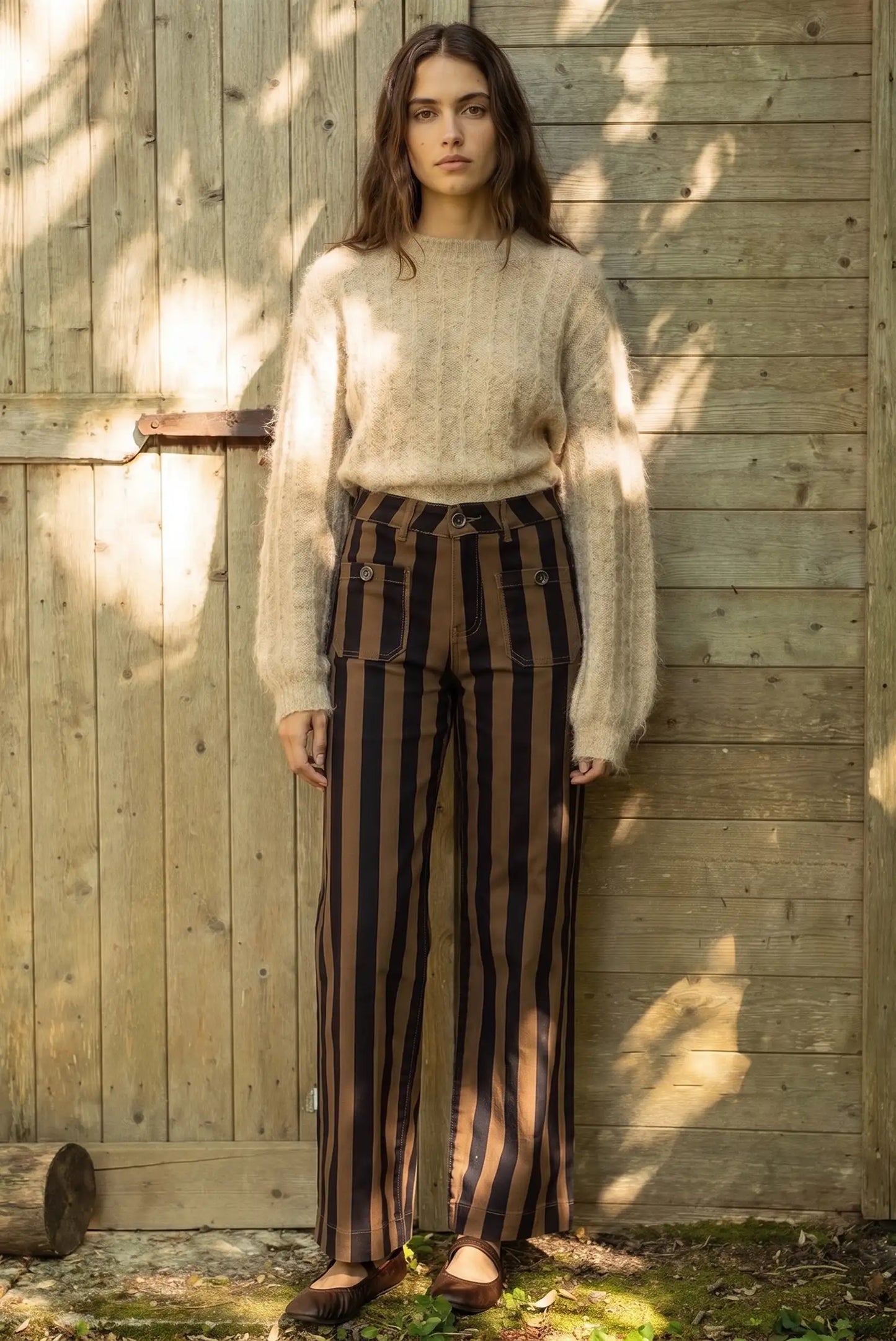 Mujer con jersey de punto beige y pantalones a rayas negras y marrones frente a pared de madera
