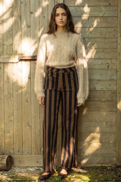 Mujer con jersey de punto beige y pantalones a rayas negras y marrones frente a pared de madera