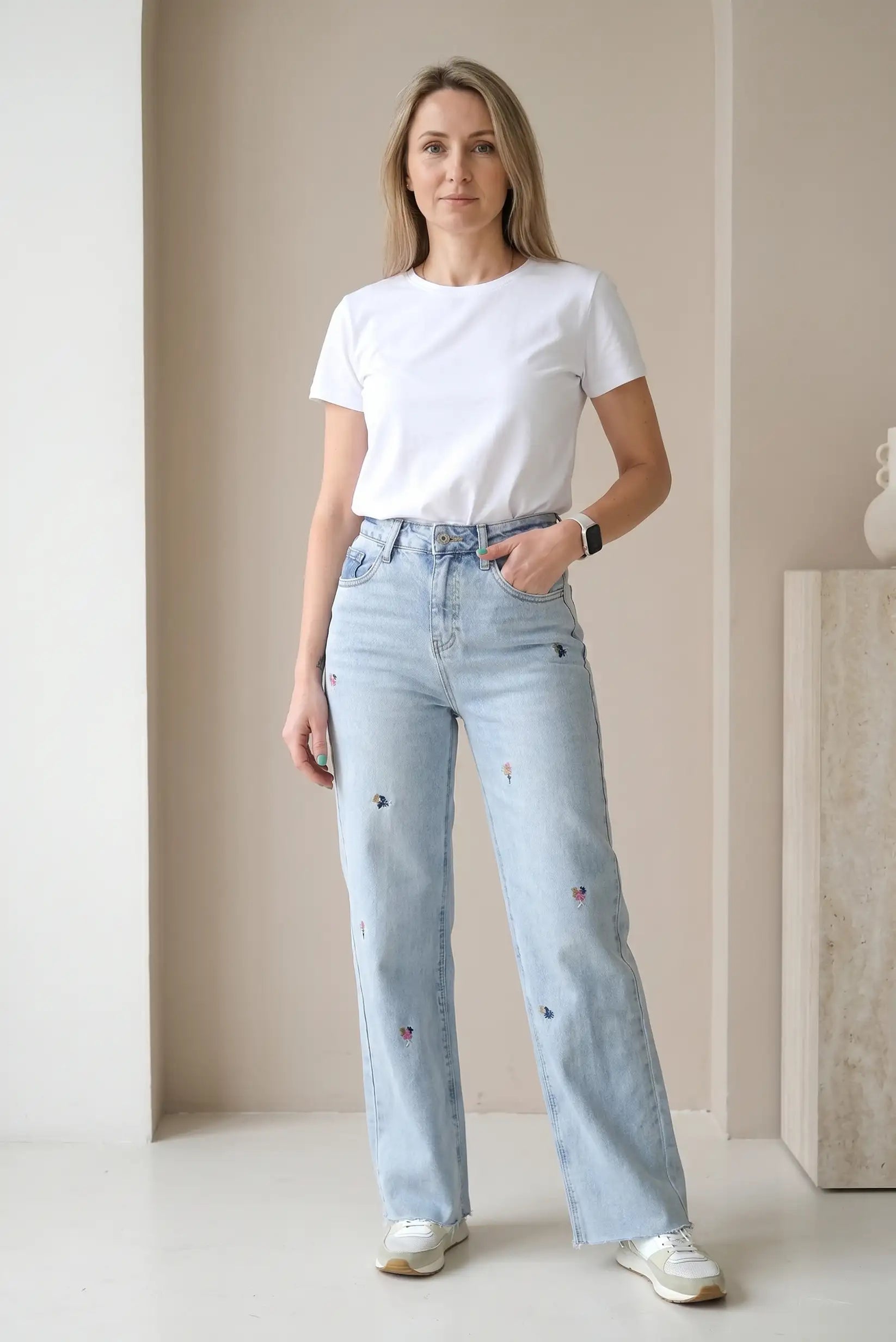 Mujer con camiseta blanca y pantalón vaquero largo azul claro con bordados de flores.