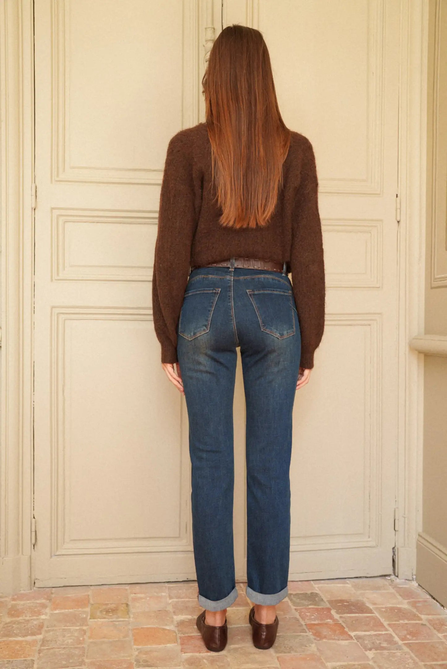 Jeans azul oscuro con bolsillos traseros, cintura alta y dobladillo enrollado, combinados con suéter marrón y zapatos marrones.