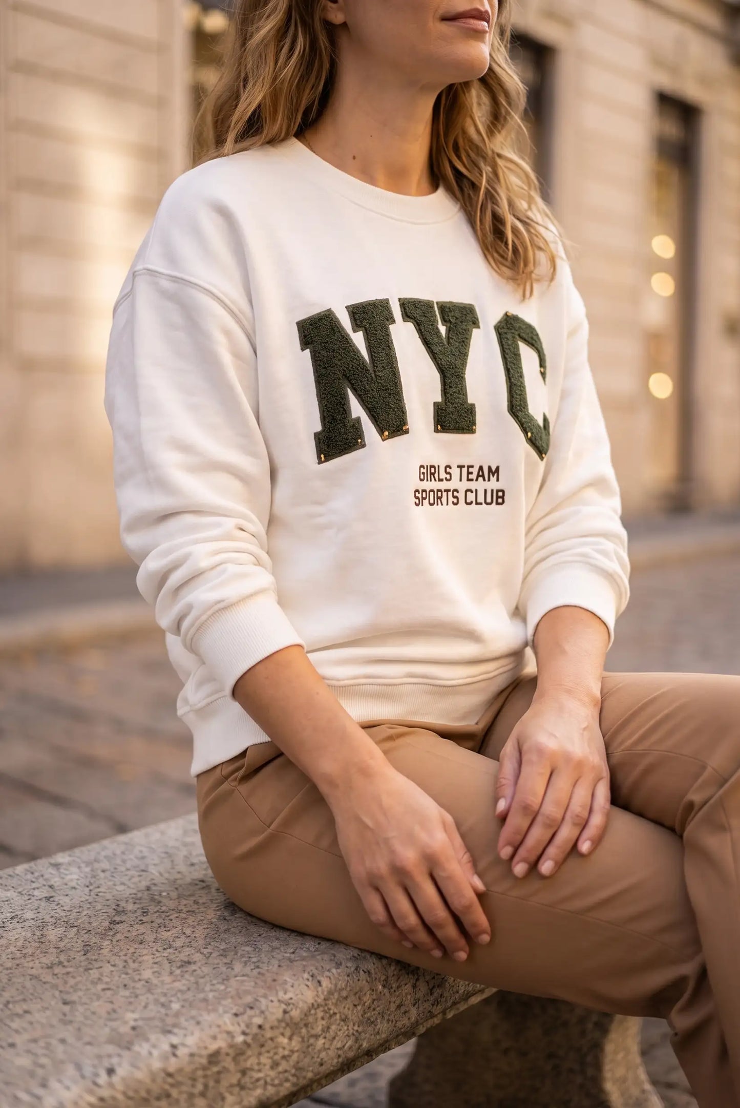Mujer vistiendo sudadera blanca con letras NYC verdes y pantalones beige.