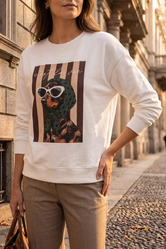 Mujer vistiendo sudadera blanca con estampado de perro con gafas y pañuelo y pantalones beige.