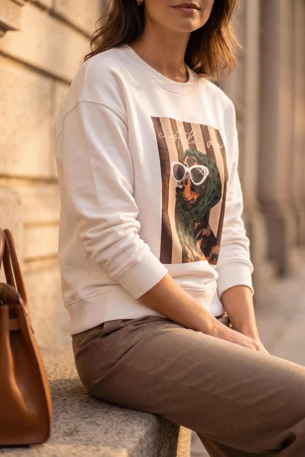 Mujer vistiendo sudadera blanca con estampado de perro con gafas de sol y pantalones marrones.