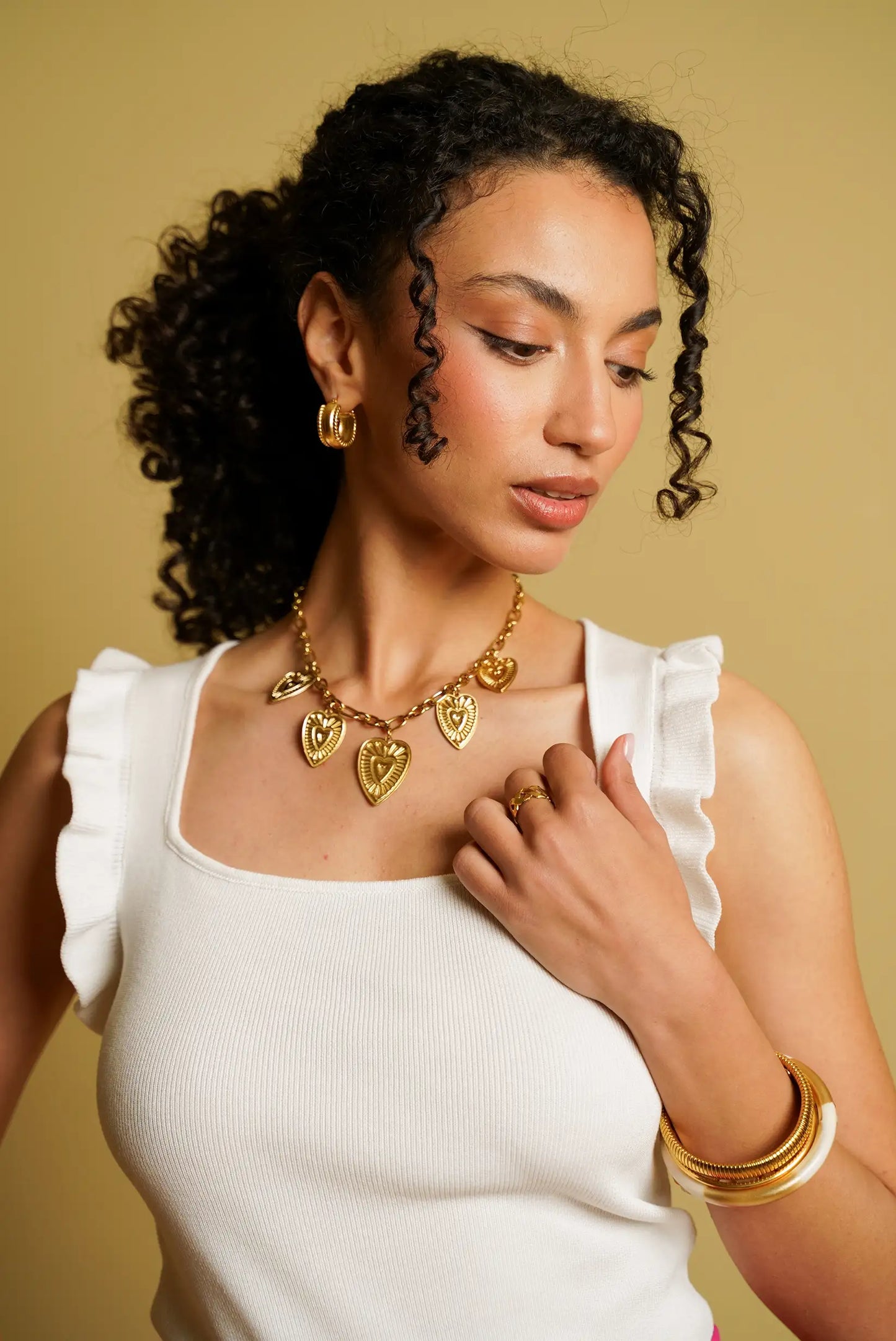 Mujer con top blanco sin mangas y volantes, collar dorado de corazones y accesorios dorados