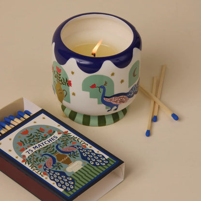 Vela decorativa Paddywax con diseño de pavo real encendida junto a caja de cerillas, en fondo beige.