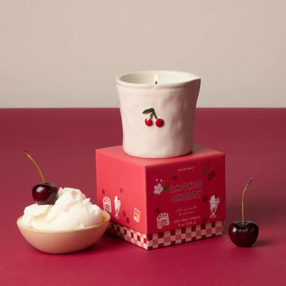 Vela aromática de cerámica blanca con cerezas, sobre caja rosa, junto a cerezas y helado.
