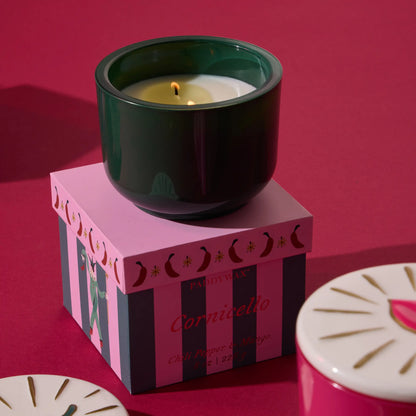 Vela Paddywax Cornicello aroma chili y mango en vaso verde sobre caja decorativa, fondo rosa.