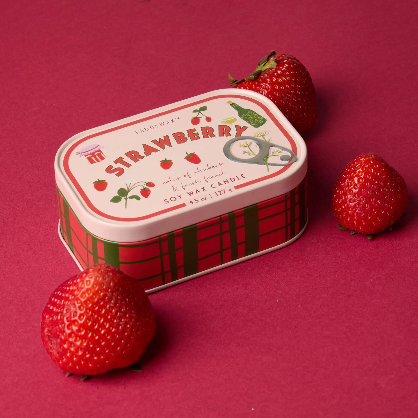 Vela Paddywax en lata roja con diseño de fresas junto a fresas frescas, sobre fondo rosa.