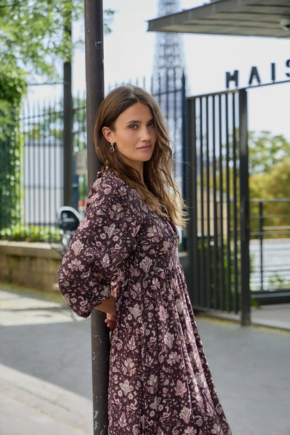 Mujer con vestido largo estampado floral en tonos burdeos apoyada en un poste metálico.