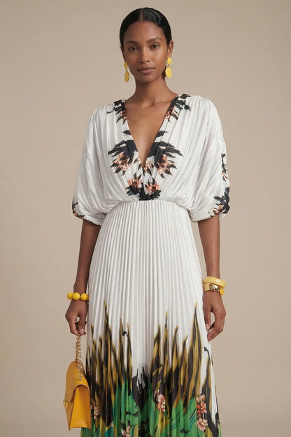 Mujer con vestido largo blanco plisado, estampado tropical verde y accesorios amarillos.