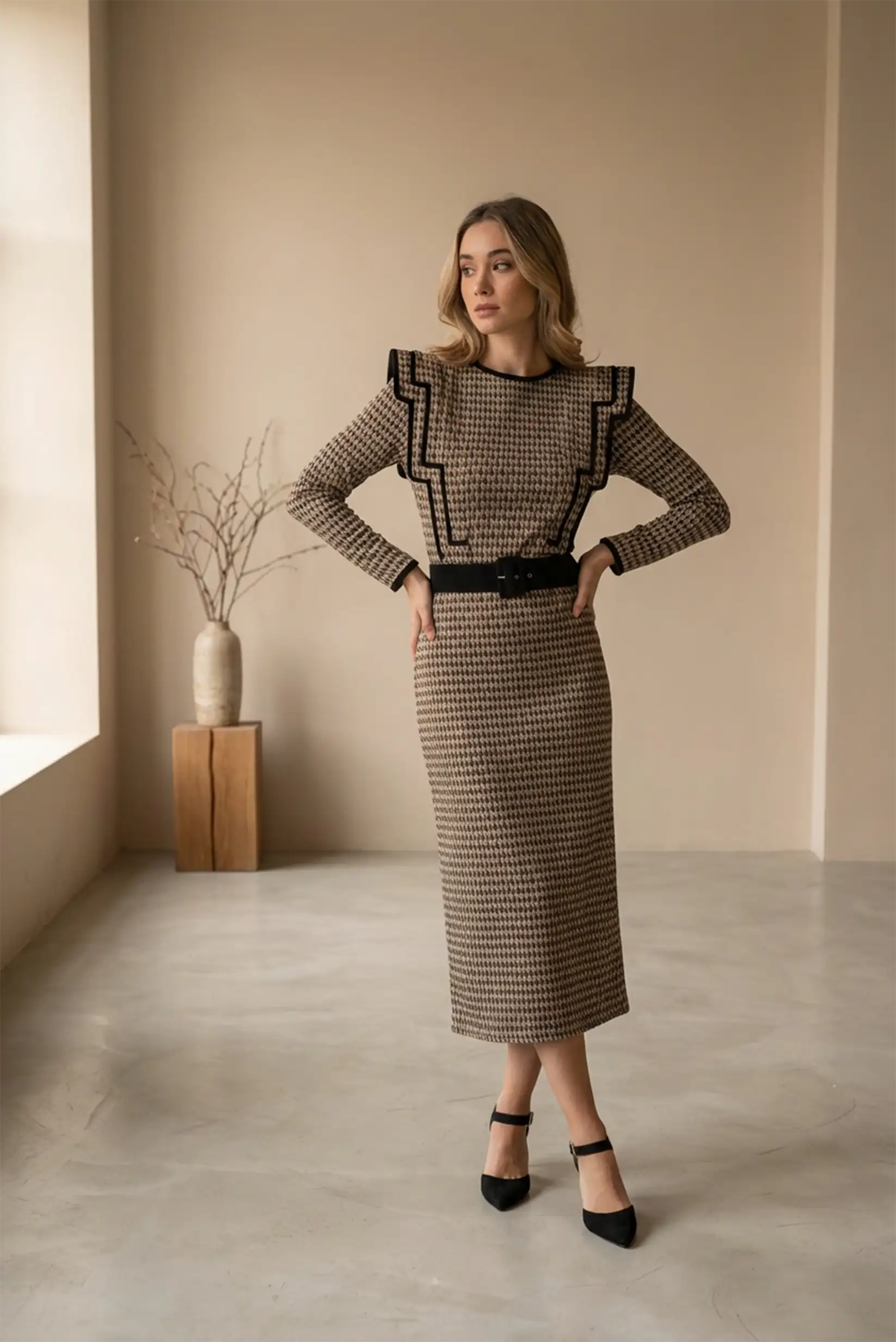 Mujer con vestido largo café clásico de manga larga y cinturón negro, en ambiente minimalista